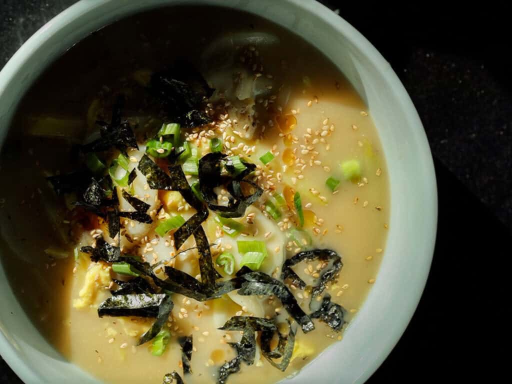 A close-up of a bowl of vegan rice cake soup in a creamy broth, topped with sliced green onions, seaweed strips, sesame seeds, and what appears to be an egg. The bowl is set against a dark background with natural lighting casting shadows.